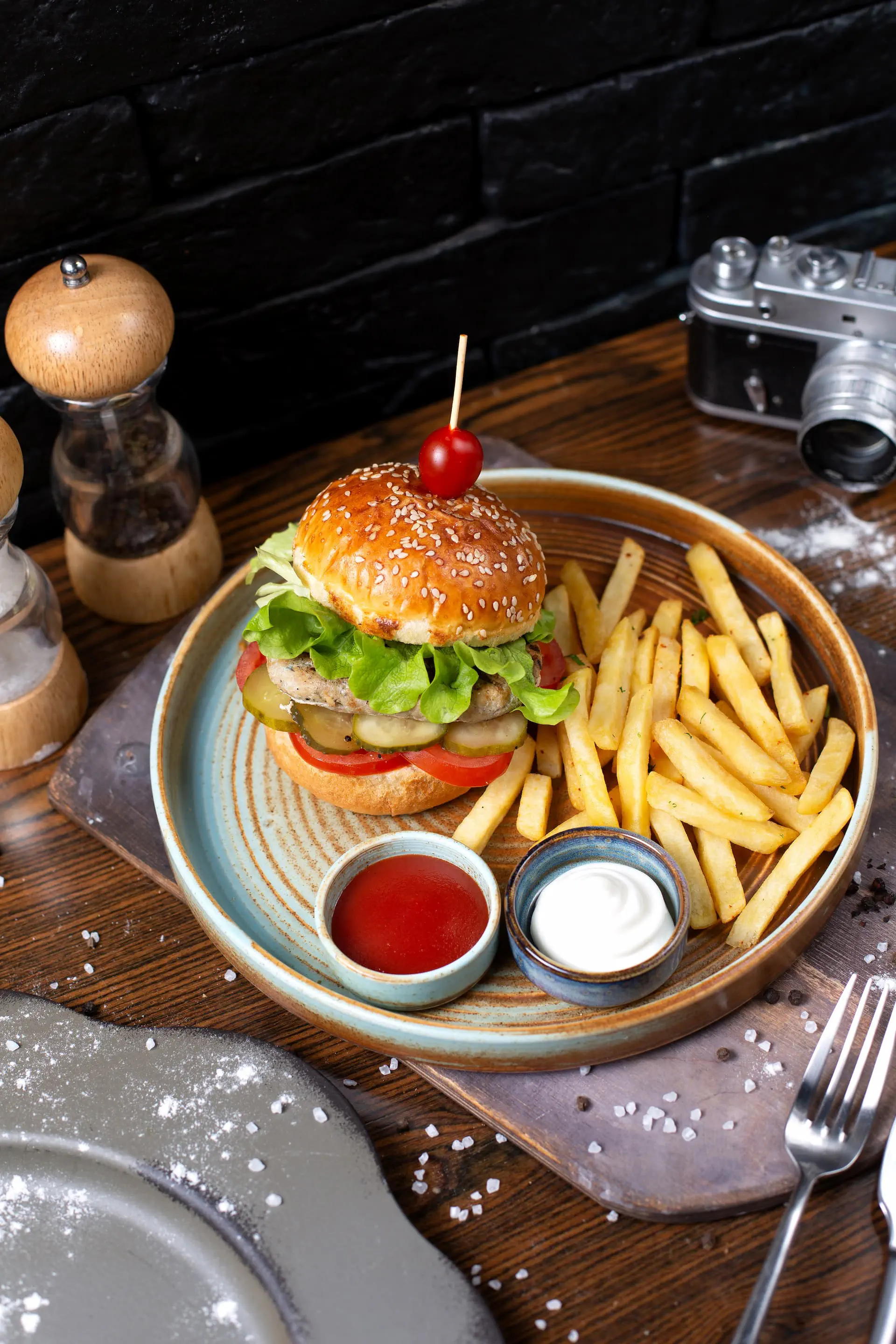 Portfolio gastronomia: Burger drobiowy z sałatą, pomidorem i ogórkiem kiszonym, podawany z frytkami i sosami na ceramicznym talerzu